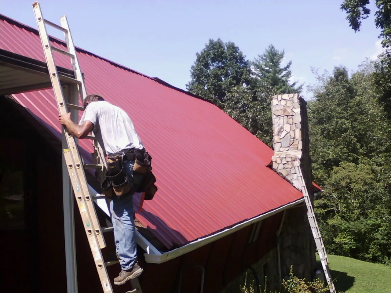 Roofer on a ladder beside a red metal roof and stone chimney — Soffit & Fascia Repair by Roofing Brothers in Cressona